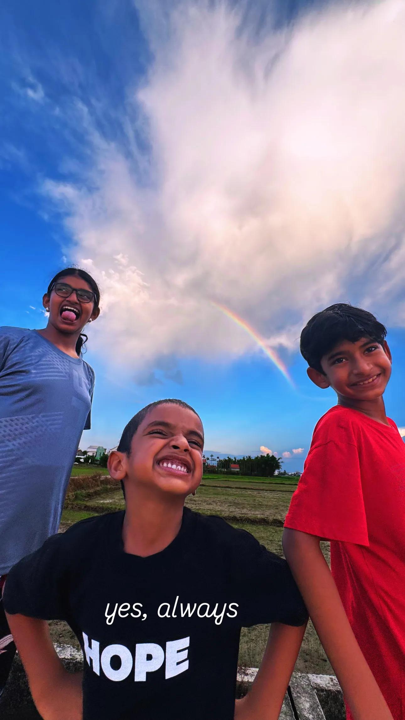 Three children laughing together outdoors under a rainbow.