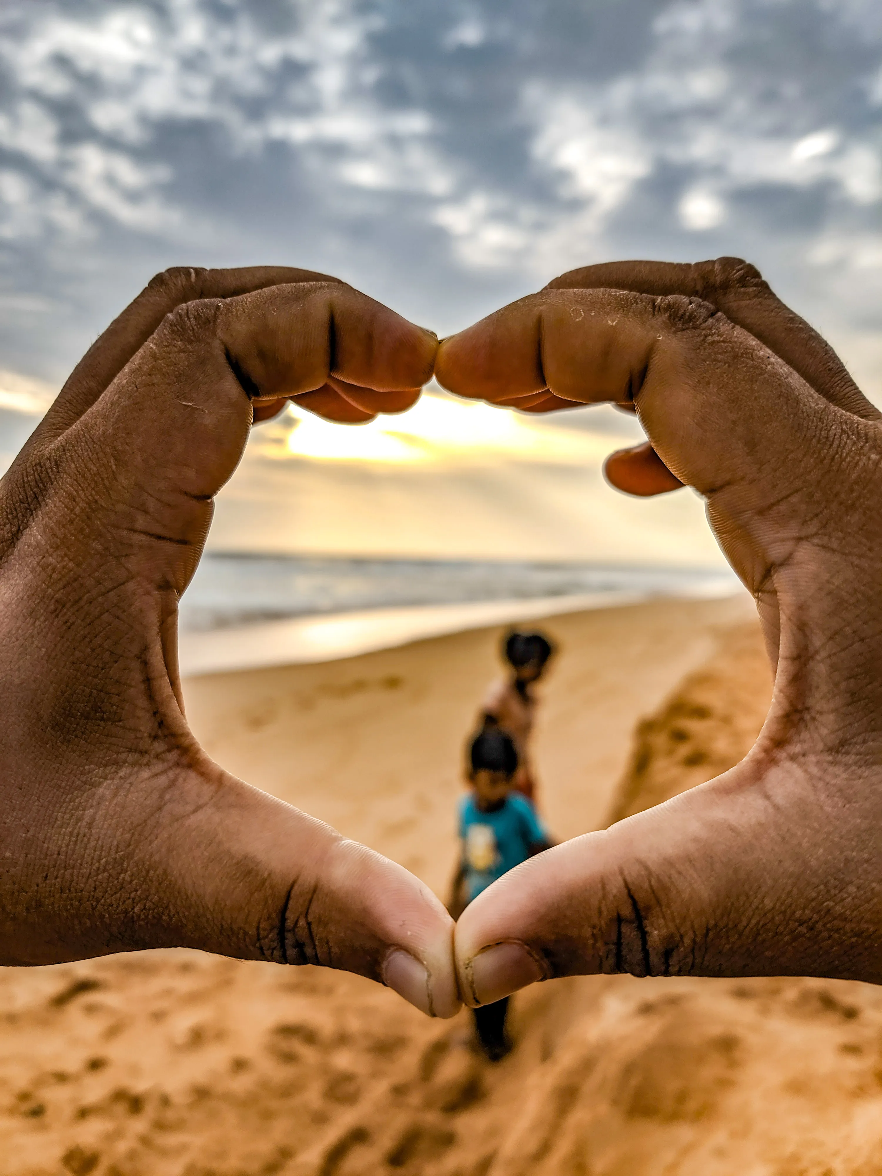 Hands forming a heart that frames two children on a beach at sunset.
