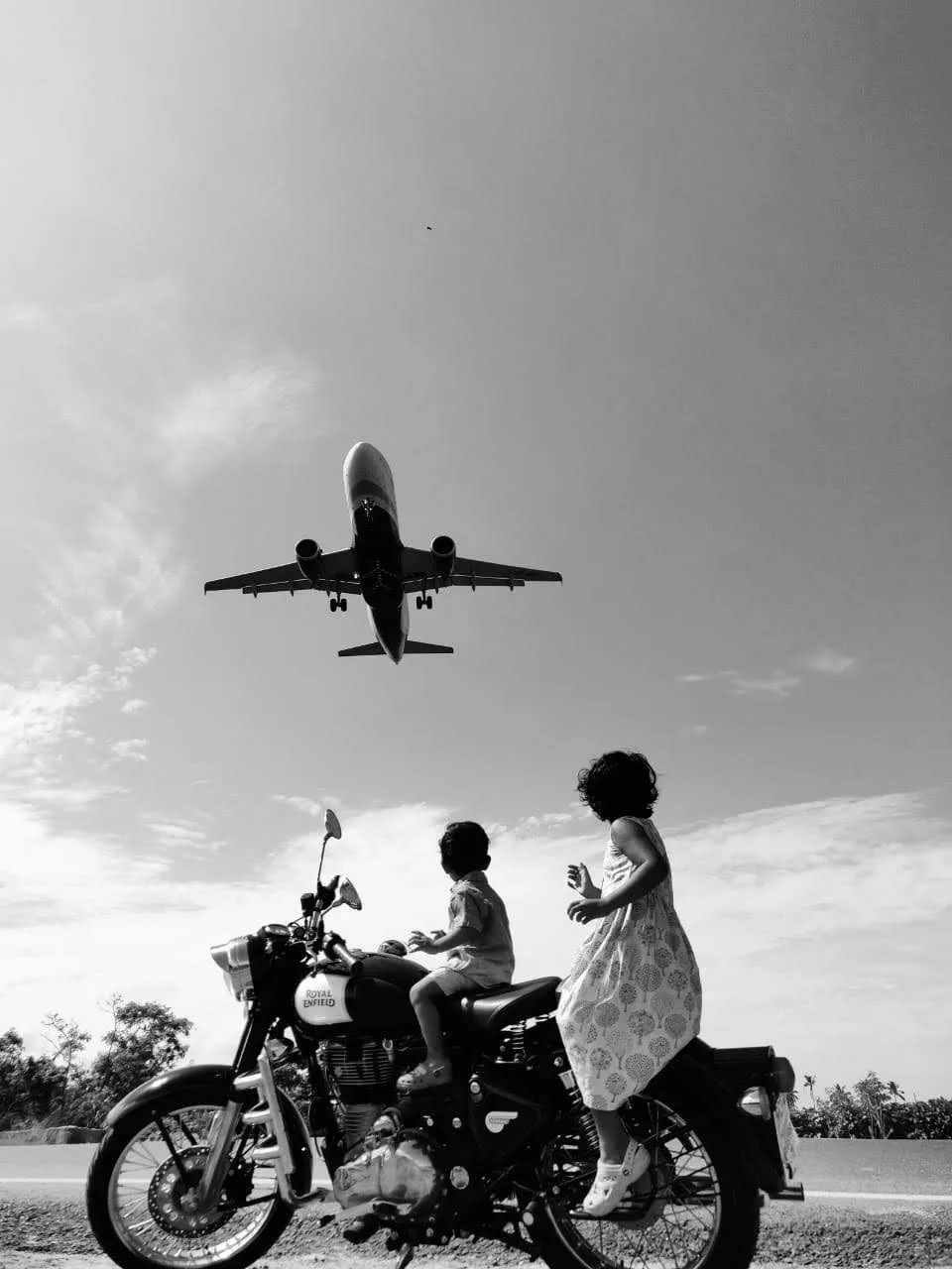Two children on a motorcycle watching a low-flying plane overhead.